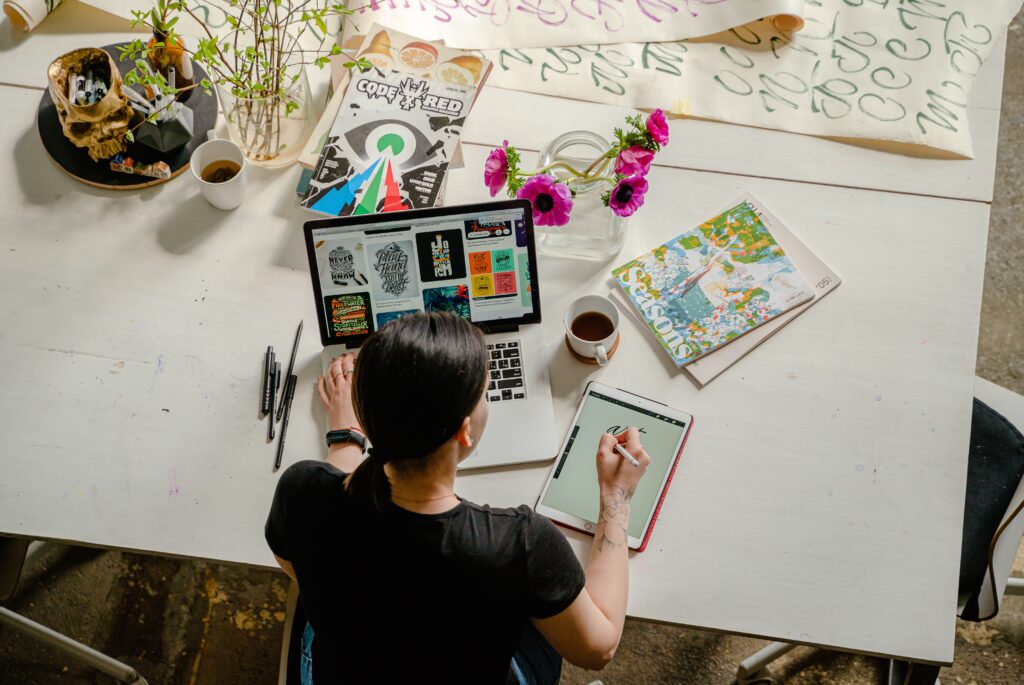 Birdseye view of lady at desk working on tablet and laptop with notepads and paper around